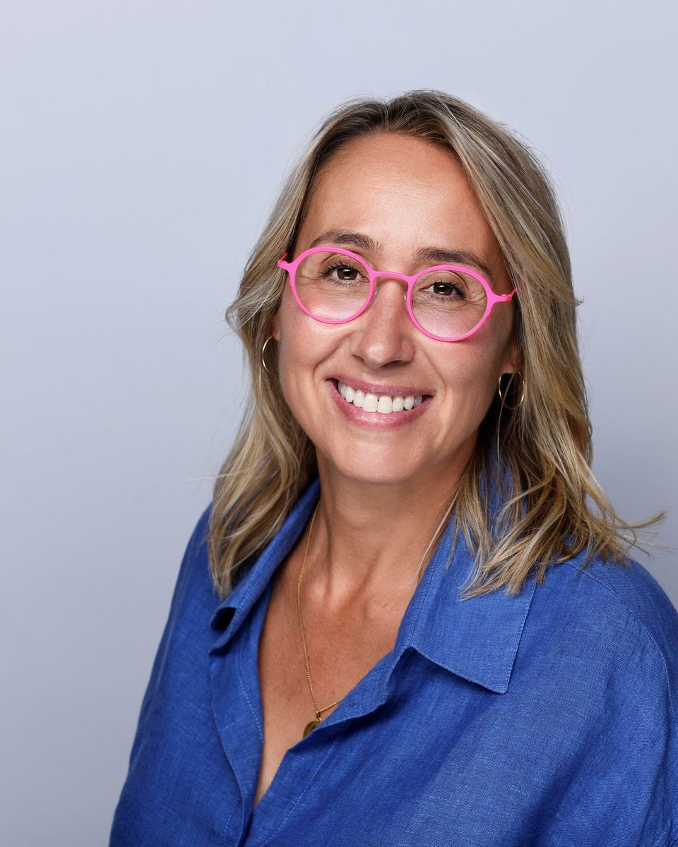 Portrait d'Agnès Rabaca, souriante, lunettes roses et chemise bleue